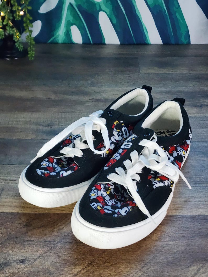 May include: A pair of black low-top sneakers featuring a repeating Mickey Mouse character print on the tongue and side panels. The shoes have solid black suede-like toe caps and heel counters, white rubber outsoles, and white lace-up closures.