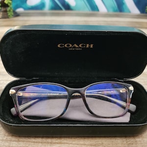 May include: A pair of dark-rimmed Coach eyeglasses with blue-tinted lenses resting inside a black hard-shell case. The interior of the case lid features the gold-colored Coach New York logo, and a gray microfiber cleaning cloth is tucked underneath.
