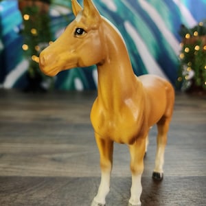 May include: A tan-colored horse figurine with a white blaze on its forehead and white lower legs. The horse is standing on a wooden surface, with a blurred background of plants and a colorful backdrop.