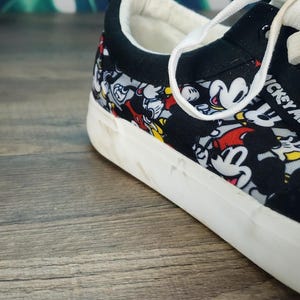 May include: A low-top sneaker featuring a black canvas upper with an all-over print of Mickey Mouse in red, yellow, and black. The shoe has a white rubber sole, white laces, and a black padded collar with a white interior lining.