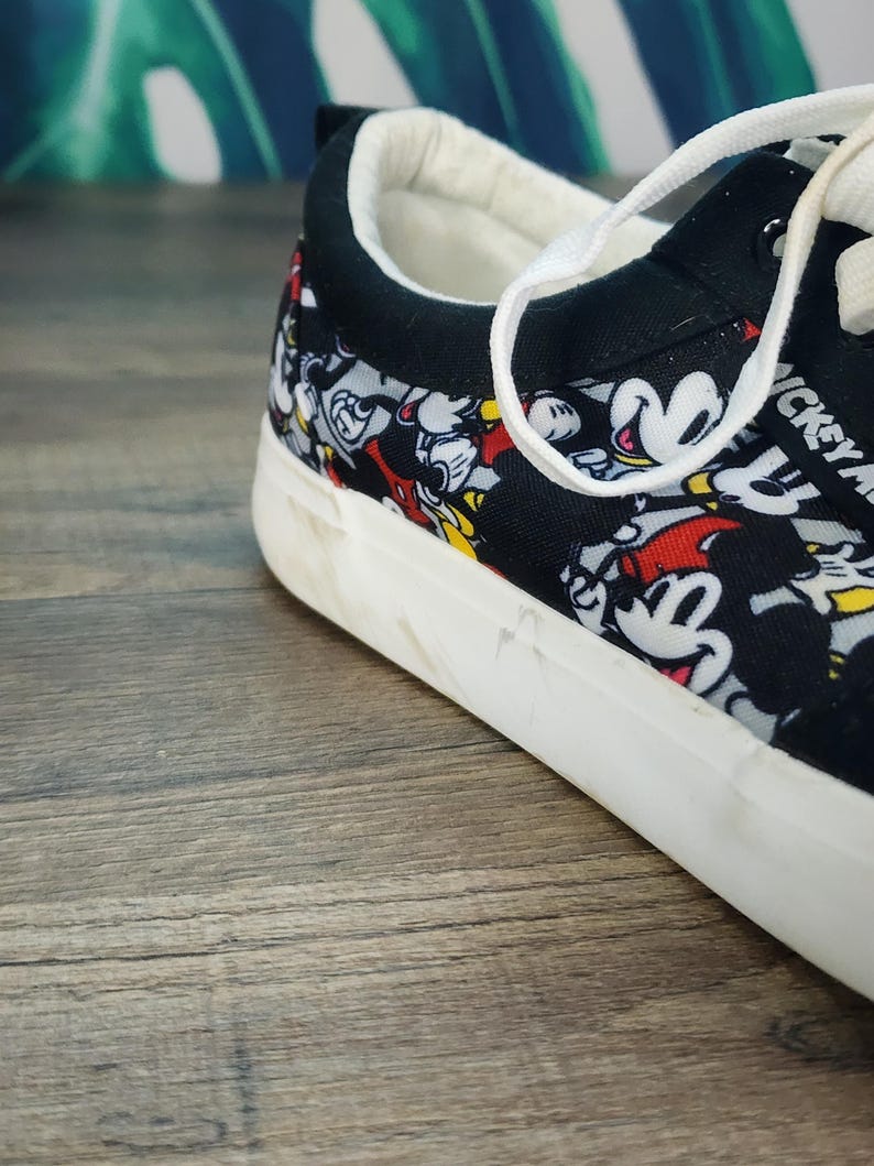 May include: A low-top sneaker featuring a black canvas upper with an all-over print of Mickey Mouse in red, yellow, and black. The shoe has a white rubber sole, white laces, and a black padded collar with a white interior lining.
