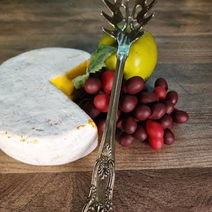Può includere: Un mestolo per pasta argentato con un manico decorativo, posizionato vicino a una fetta di formaggio, una mela verde e uva rossa. La testa del mestolo ha un design a stella. La composizione è su una superficie di legno.