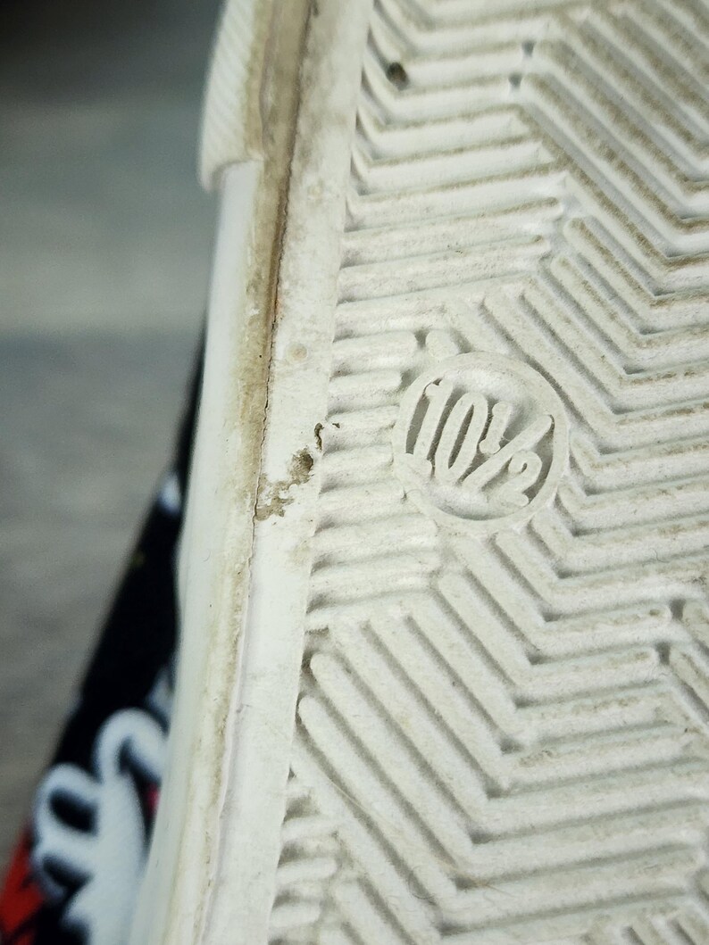 May include: The white rubber sole of a shoe features a textured herringbone tread pattern and a circular embossed marking displaying the size 10.5. The edge of the sole shows signs of wear, and a portion of the black patterned upper is visible.