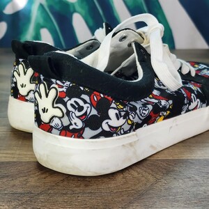 May include: Low-top sneakers featuring a repeating pattern of Mickey Mouse faces and white gloved hands on a black, red, and grey background. The shoes have black fabric trim, white laces, and a thick white rubber sole with a textured finish.