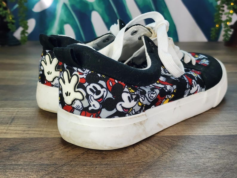 May include: Low-top sneakers featuring a repeating pattern of Mickey Mouse faces and white gloved hands on a black, red, and grey background. The shoes have black fabric trim, white laces, and a thick white rubber sole with a textured finish.