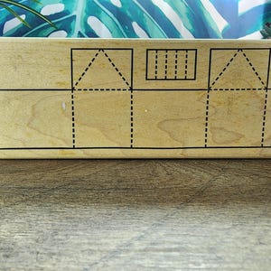 May include: A rectangular wooden stamp with a house design. The stamp features black outlines and dashed lines indicating where to fold. The design includes a roof, walls, and a chimney, perfect for crafting projects.