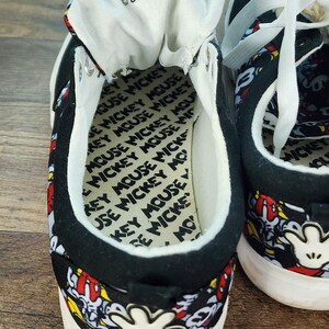 May include: A pair of black low-top sneakers featuring an all-over Mickey Mouse print with white laces and white soles. The interior insole is printed with the text Mickey Mouse repeatedly, and the tongue features the Disney logo in black text.