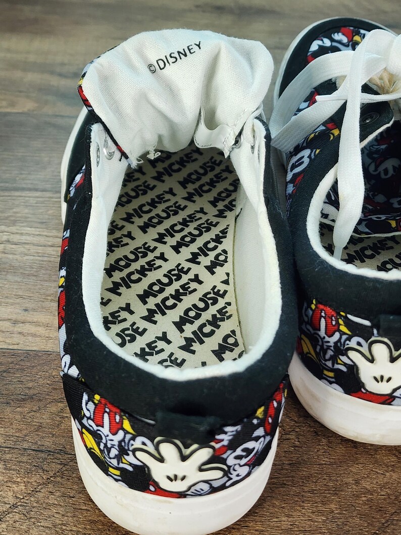 May include: A pair of black low-top sneakers featuring an all-over Mickey Mouse print with white laces and white soles. The interior insole is printed with the text Mickey Mouse repeatedly, and the tongue features the Disney logo in black text.