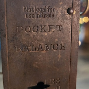 May include: Close-up of a vintage pocket balance scale with the text "Not legal for use in trade." The scale is made of aged metal with the word "POCKET BALANCE" embossed on the front. The scale reads in pounds (lbs).