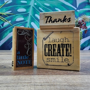 Puede incluir: Dos sellos de goma de madera rectangulares. El sello vertical más pequeño presenta un panel oscuro con el texto 'a little NOTE' y remolinos decorativos. El sello cuadrado más grande presenta el texto 'Thanks' sobre 'laugh. CREATE! smile.' dentro de un borde de pergamino.