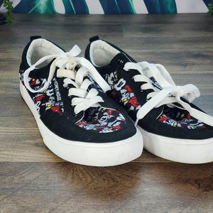 May include: A pair of black low-top sneakers featuring a repeating Mickey Mouse graphic print in red, white, and black. The shoes have white laces, white rubber soles, and the text Mickey Mouse printed in white along the side of the eyelet panel.
