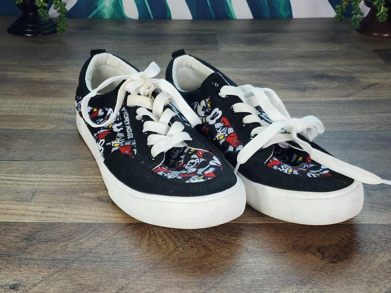 May include: A pair of black low-top sneakers featuring a repeating Mickey Mouse graphic print in red, white, and black. The shoes have white laces, white rubber soles, and the text Mickey Mouse printed in white along the side of the eyelet panel.