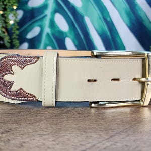 May include: Cream-colored leather belt with a decorative brown and tan design. The belt features a gold-colored buckle and a stitched design. A stylish accessory for any outfit.