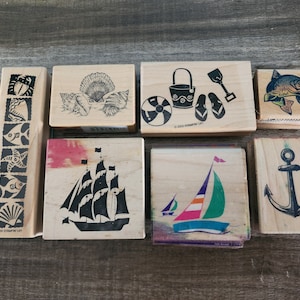 以下が含まれることがあります： ビーチと航海のテーマの木製スタンプのコレクション。デザインには、貝殻、ヨット、錨、魚、ビーチアクセサリーが含まれます。スタンプは木製で、黒インクのデザインが施されています。