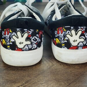 May include: A pair of low-top sneakers featuring a black fabric upper with a repeating pattern of Mickey Mouse faces in red, white, and black. Each heel displays a prominent white cartoon glove graphic, white laces, and a thick white rubber sole.