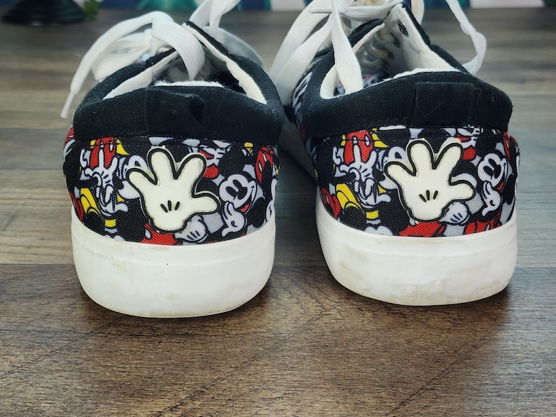 May include: A pair of low-top sneakers featuring a black fabric upper with a repeating pattern of Mickey Mouse faces in red, white, and black. Each heel displays a prominent white cartoon glove graphic, white laces, and a thick white rubber sole.
