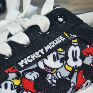 May include: A black canvas sneaker featuring a repeating pattern of Mickey Mouse in red, yellow, and white. The shoe has white laces, black metal eyelets, and a black fabric tag with the white text Mickey Mouse stitched onto the side panel.