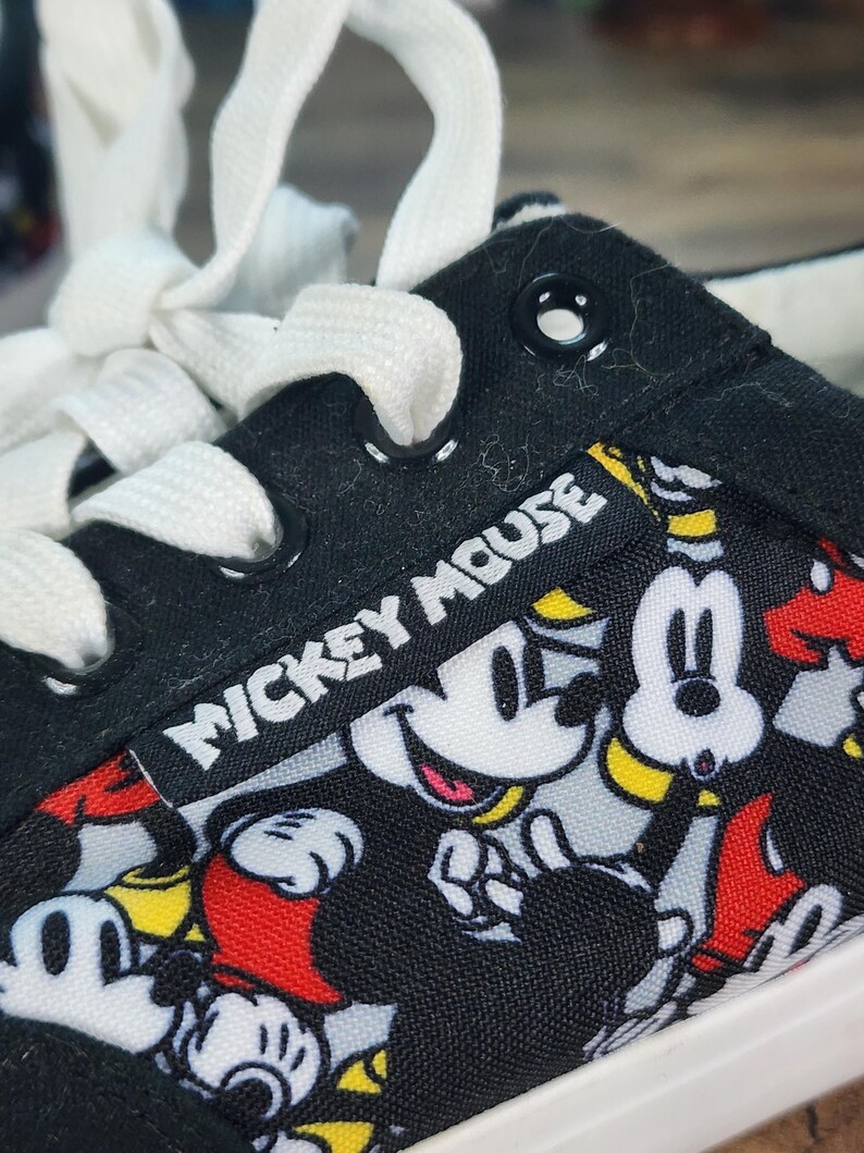 May include: A black canvas sneaker featuring a repeating pattern of Mickey Mouse in red, yellow, and white. The shoe has white laces, black metal eyelets, and a black fabric tag with the white text Mickey Mouse stitched onto the side panel.