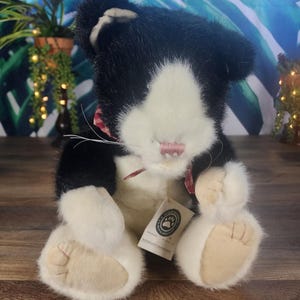 May include: A plush black and white cat stuffed animal with a pink nose and a red and white checkered ribbon. The cat has white paws and a tag attached to its ear.