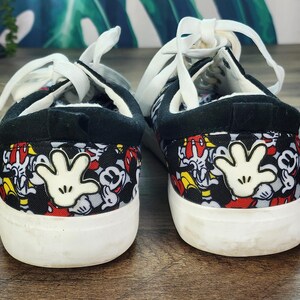 May include: A pair of low-top sneakers featuring a black fabric upper with an all-over print of Mickey Mouse faces and a prominent white cartoon glove graphic on the heel. The shoes have white laces, a black padded collar, and a thick white rubber sole.