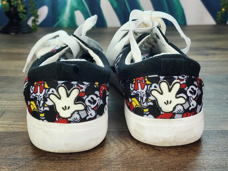 May include: A pair of low-top sneakers featuring a black fabric upper with an all-over print of Mickey Mouse faces and a prominent white cartoon glove graphic on the heel. The shoes have white laces, a black padded collar, and a thick white rubber sole.