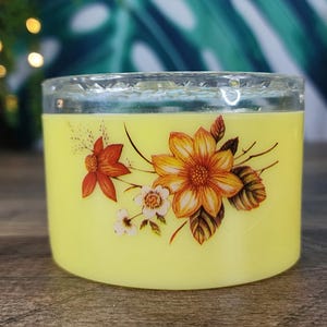 Pode incluir: Uma vela amarela cilíndrica em um castiçal de vidro transparente. A vela é decorada com um desenho floral com flores laranja, amarelas e brancas com folhas marrons. O castiçal tem uma borda decorativa facetada.