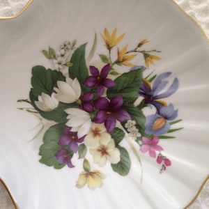 May include: A white porcelain shell-shaped dish with a gold rim. The dish features a hand-painted floral design with purple, white, yellow, and blue flowers.