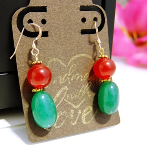 May include: A pair of dangle earrings featuring a red, round bead above a green, oval bead. The earrings are suspended from gold-colored hooks and are displayed on a brown card with the words "handmade with love".