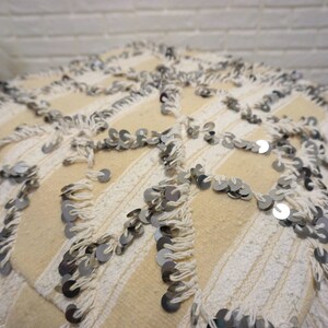 May include: A cream-colored throw pillow with white fringe and silver sequins. The pillow has a geometric pattern.