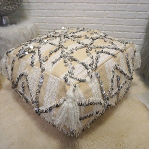 May include: A cream-colored pouf with a geometric pattern and silver sequins. The pouf has white tassels around the edges.