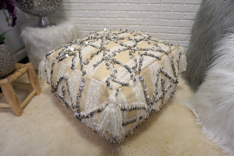 May include: A cream-colored pouf with a geometric pattern and silver sequins. The pouf has white tassels around the edges.
