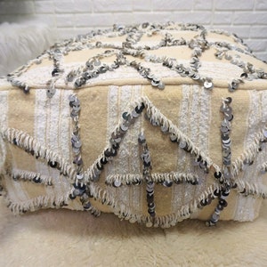 May include: A cream-colored pouf with a geometric pattern and silver sequins. The pouf has white fringe around the edges and white tassels on the sides.