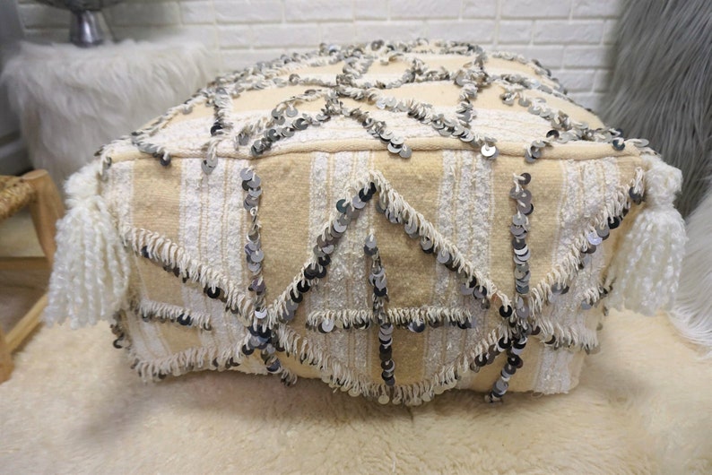 May include: A cream-colored pouf with a geometric pattern and silver sequins. The pouf has white fringe around the edges and white tassels on the sides.
