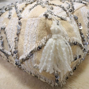 May include: A white floor cushion with a fringe border and silver sequins. The cushion has a white tassel on one side.