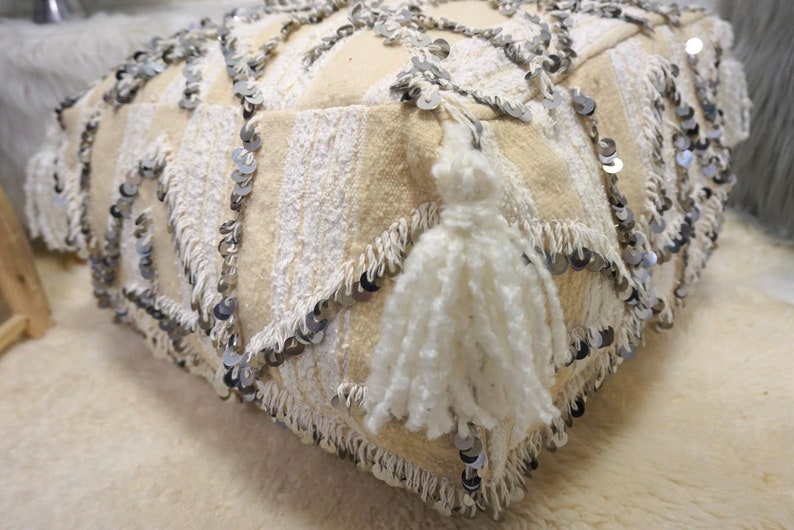 May include: A white floor cushion with a fringe border and silver sequins. The cushion has a white tassel on one side.