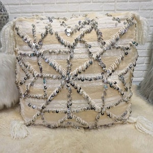 May include: A large, square, off-white floor cushion with a geometric pattern of silver sequins and fringe. The cushion has tassels on each corner.