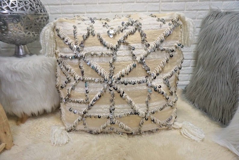 May include: A large, square, off-white floor cushion with a geometric pattern of silver sequins and fringe. The cushion has tassels on each corner.