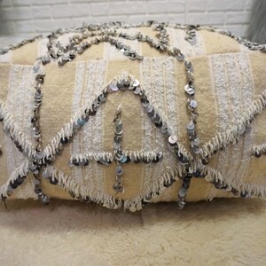 May include: A cream-colored pouf with white fringe and silver sequins. The pouf is made of a soft, textured fabric and has a unique, bohemian style.