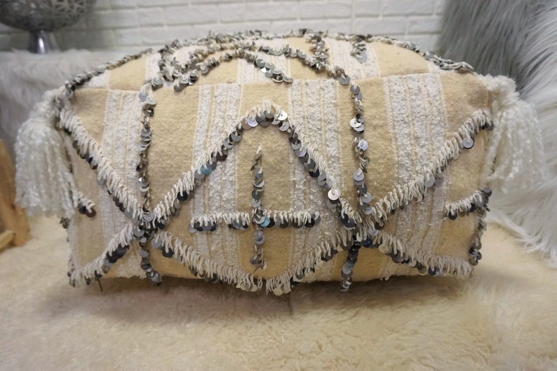 May include: A cream-colored pouf with white fringe and silver sequins. The pouf is made of a soft, textured fabric and has a unique, bohemian style.