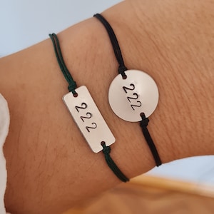 May include: Two bracelets with black and green cords. One bracelet has a round silver charm with the number 222 engraved on it. The other bracelet has a rectangular silver charm with the number 222 engraved on it.