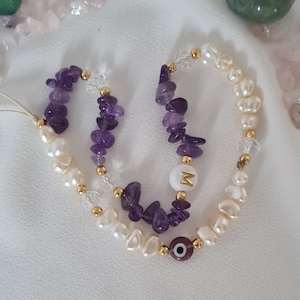 Puede incluir: Un brazalete de amatista púrpura y perlas blancas con un dije de oro que dice "M" y un dije azul de ojo turco.
