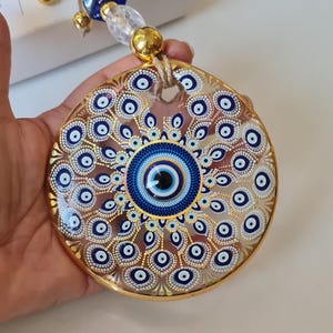 May include: A round, gold-rimmed Evil Eye charm with a blue and white eye design. The charm is suspended by a natural twine cord, with clear and gold beads. The charm is held in a hand.