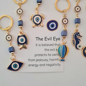 Puede incluir: Llaveros dorados con amuletos de ojo turco azules y blancos. Los amuletos incluyen un pez, una tortuga, un caballito de mar y un globo aerostático. El texto en la tarjeta dice "The Evil Eye" y explica sus propiedades protectoras.