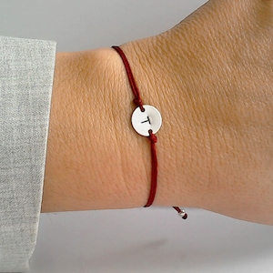 May include: A red string bracelet with a silver charm featuring the letter "T".