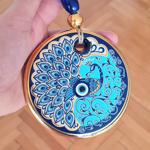 Peut inclure: Une décoration en céramique bleue et dorée avec un motif de paon et un mauvais œil bleu. La décoration est suspendue à une chaîne de perles bleues et dorées.