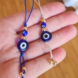 Puede incluir: Dos llaveros con amuleto de ojo turco azul con cuentas doradas y una cuerda azul. Los amuletos son redondos y tienen un ojo blanco en el centro.