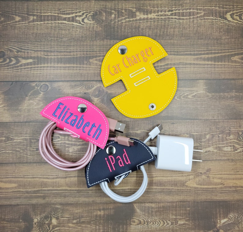 May include: Three cord organizers made of leather with embroidered names. The organizers are yellow, pink, and black. The yellow organizer says "Car Charger", the pink organizer says "Elizabeth", and the black organizer says "iPad".