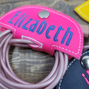 May include: Pink leather cord organizer with a snap closure and the name "Elizabeth" embroidered in blue thread.