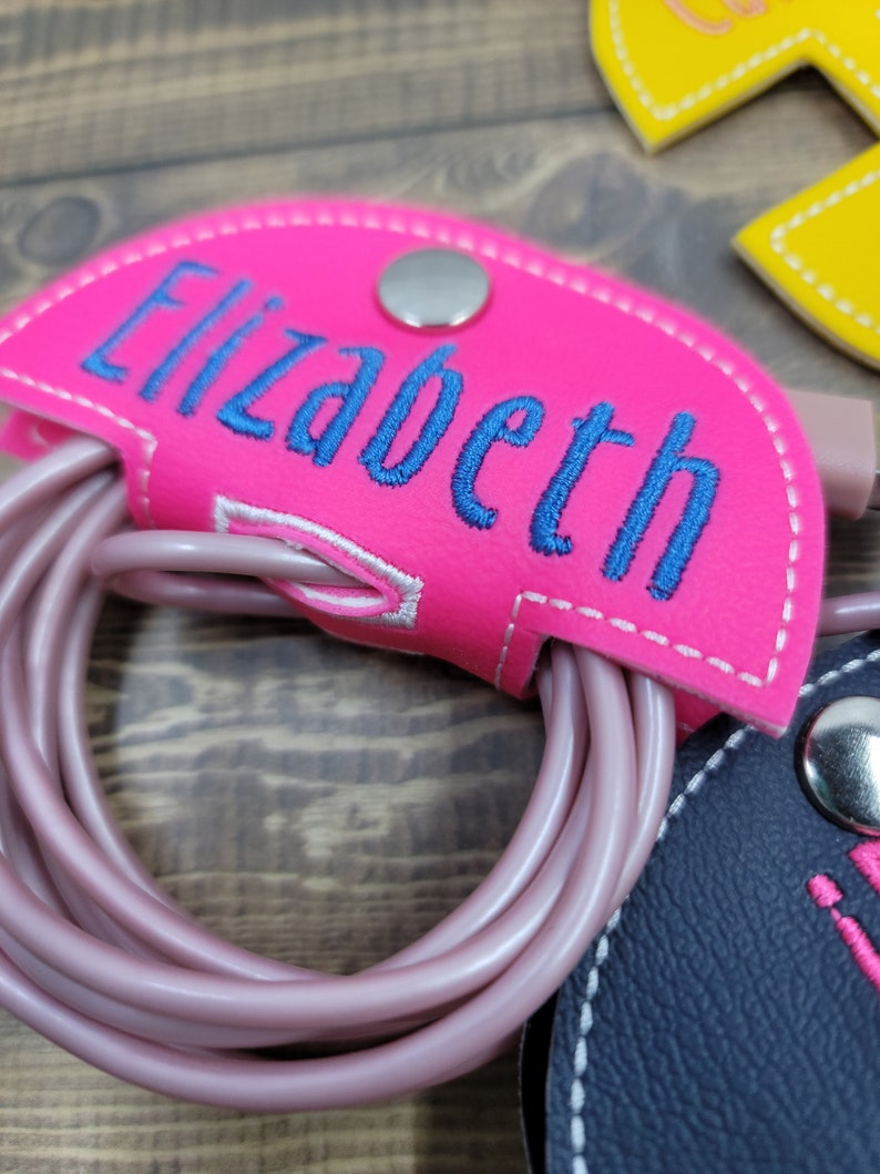 May include: Pink leather cord organizer with a snap closure and the name "Elizabeth" embroidered in blue thread.