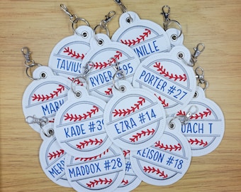 May include: A collection of white and grey baseball-themed keyrings with red stitching. Each keyring features a different name and number, such as "TAVIUS", "RYDER 95", "PORTER #27", "KADE #3", "EZRA #14", "KEISON #18", "MADDOX #28", and "COACH T".